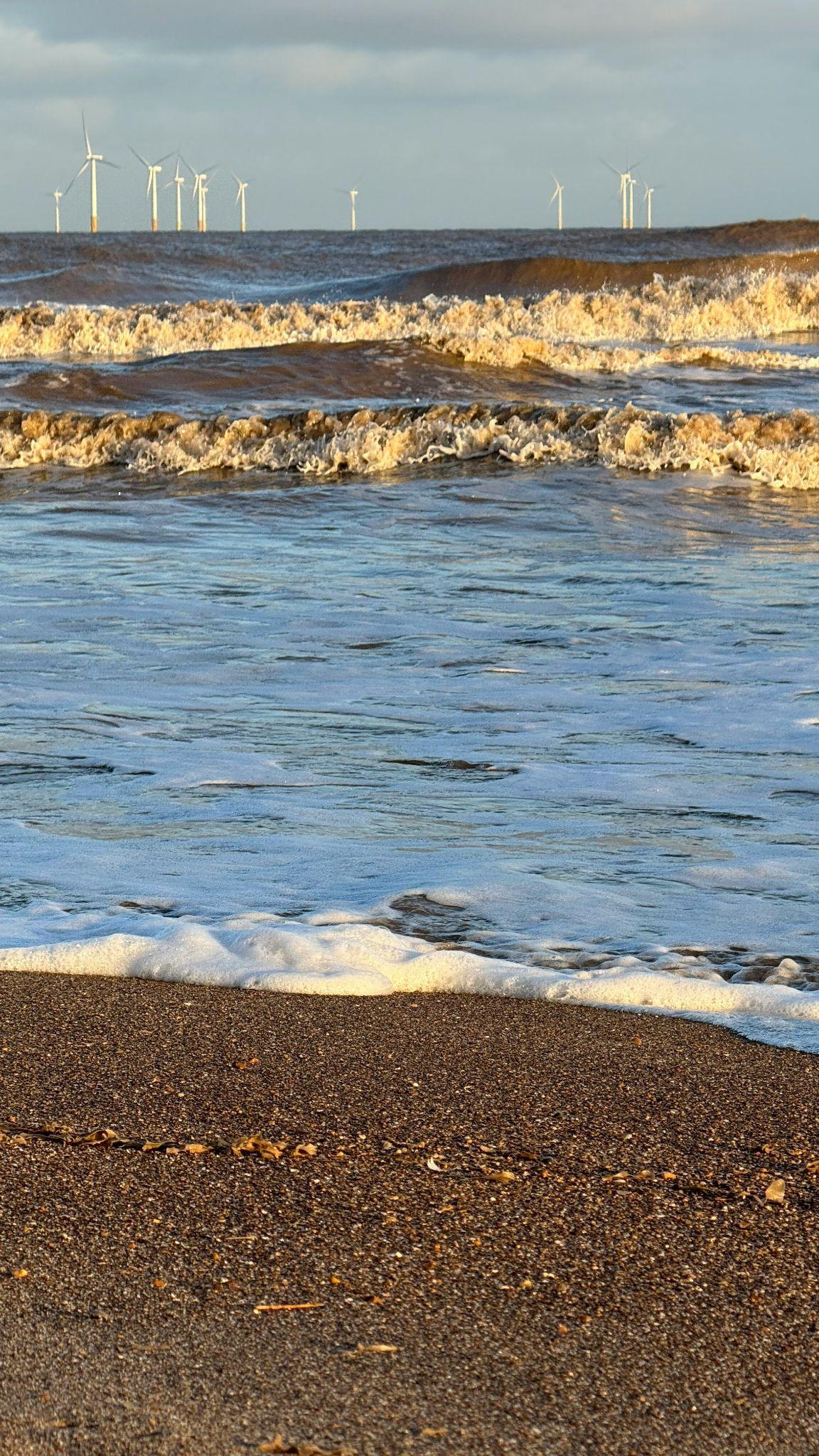 Waves breaking on the sand at Saltfleet beach at golden hour, with offshore wind turbines on the horizon